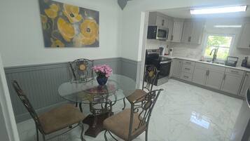 A kitchen with white cabinets, a glass dining table with chairs, a painting on the wall, and a window with a view.