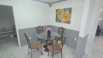 A dining area with a glass table, four chairs, a vase with flowers, and a painting on the wall.