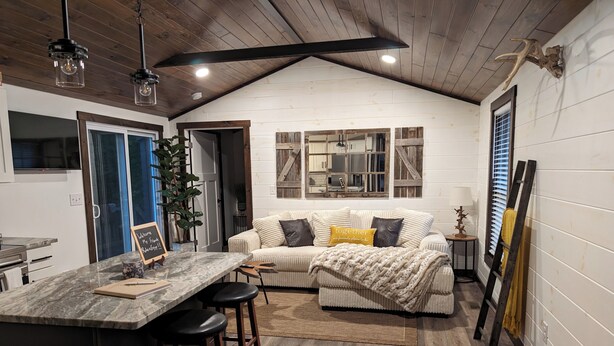 A cozy kitchen with a wooden ceiling, a bar area with stools, a sofa with cushions, and a rustic decor including a deer head on the wall. - Horse-Friendly Retreat:Trail Access, Woodland Views & Peaceful Deck (Allardt)