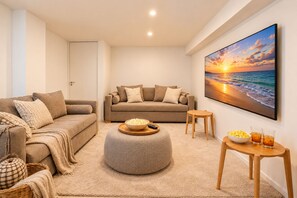 A modern living room with a beige sofa, a round ottoman, a small wooden table with snacks, and a large painting of a sunset over the ocean. - Signature Lake View Luxury Residences Unite Ñ36 (San Carlos de Bariloche)