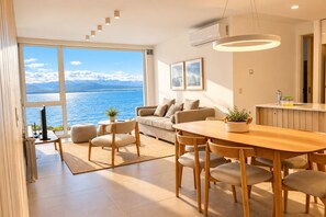 A modern living space with a large window overlooking a body of water, a dining area with wooden furniture, and a kitchen area with a built-in counter. - Signature Lake View Luxury Residences Unite Ñ36 (San Carlos de Bariloche)