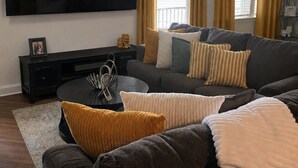 Ein Wohnzimmer mit großem Flachbildfernseher, einer Couch, einem Couchtisch und gelben Vorhängen.