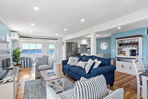 A modern living room with a blue sofa, striped armchairs, a wooden coffee table, and a view of the ocean through large windows. - Waterfront Cottage (Swansea)