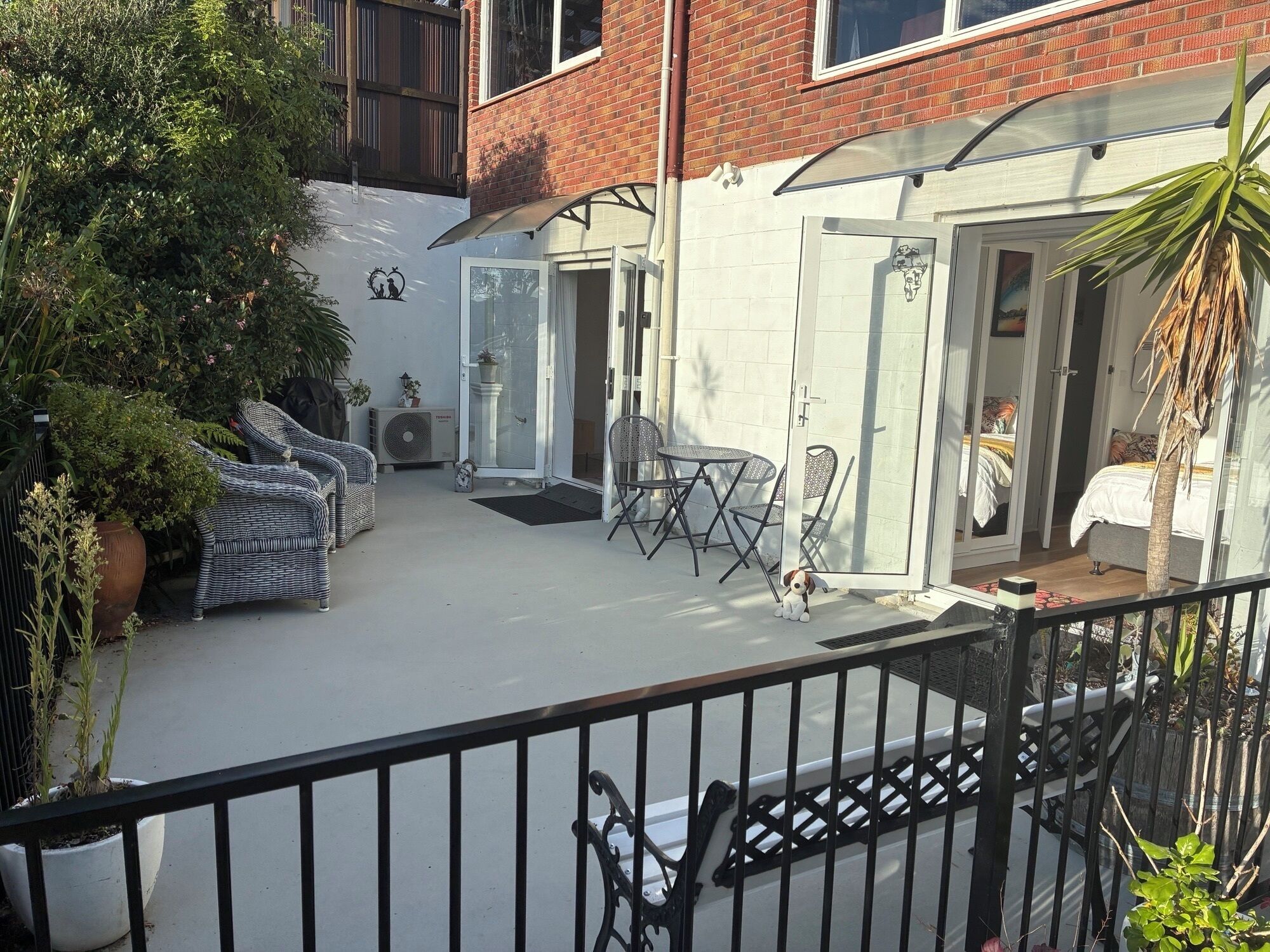 Eine Terrasse mit Gartenmöbeln, einem Hund und Blick auf ein Schlafzimmer durch eine Glastür.
