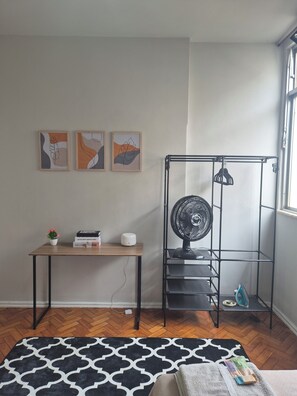 Ein Zimmer mit einem Bett, einem Schreibtisch mit Topfpflanze, einem Standventilator und einem Metallregal.