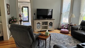 Ett vardagsrum med en tv, en grå fåtölj, en svart lädersoffa, ett träbord och en mönstrad matta.
