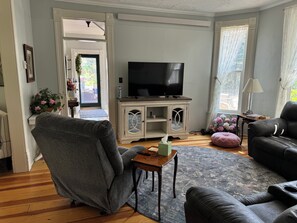 A living room with a television, a grey armchair, a black leather sofa, a wooden coffee table, and a patterned rug. -  Beautiful Victorian style home in a quiet serene area but close to attractions (Lead)