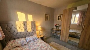 A bedroom with a tufted headboard, a floral bedspread, a bedside lamp, a small table with a vase of flowers, and a mirror on the door.