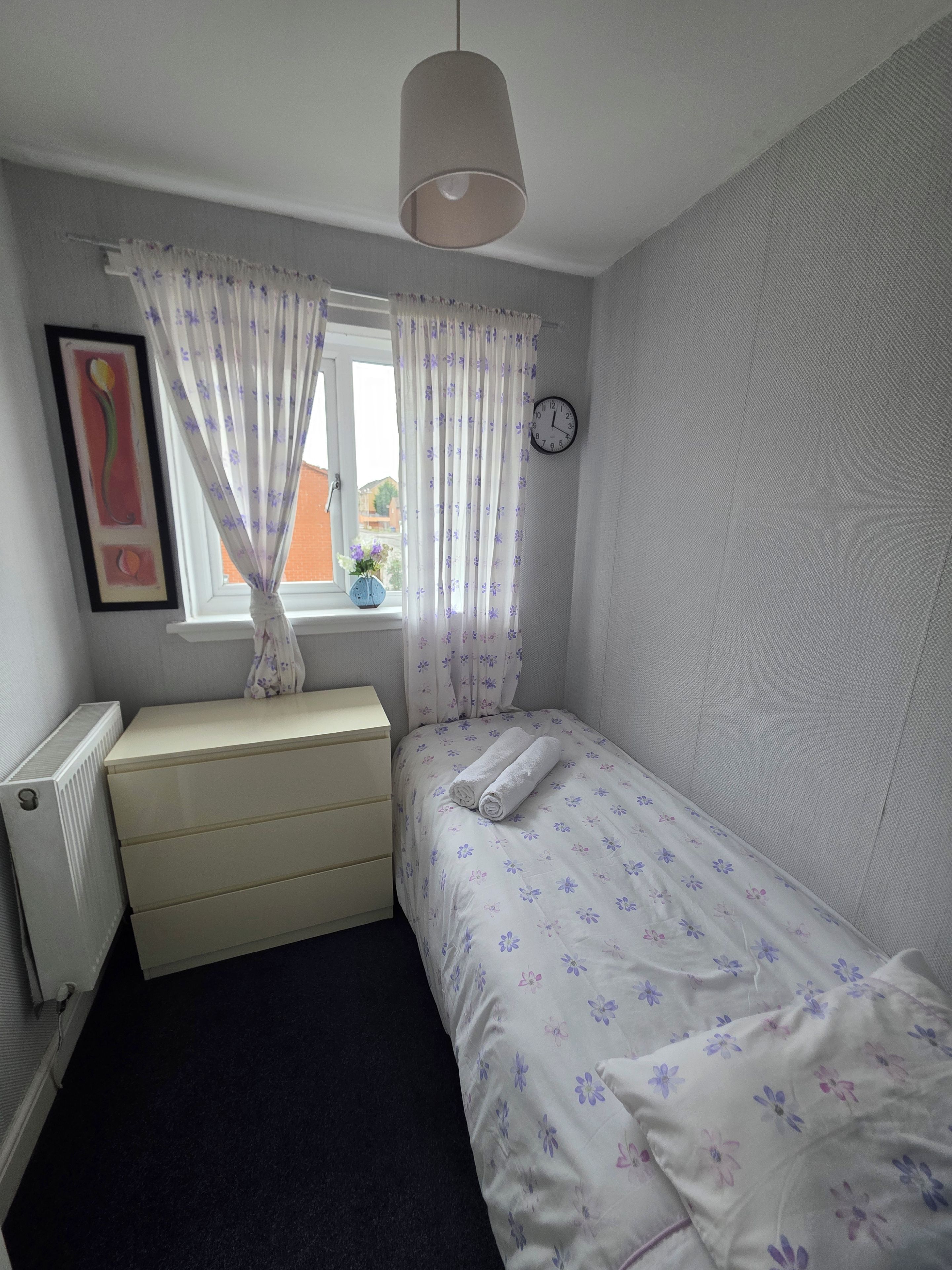 A bedroom with a single bed, a dresser, a radiator, a window with curtains, a clock, and a framed picture on the wall.