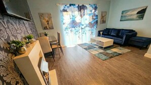 A living room with a sofa, coffee table, and chairs, a TV mounted on the wall, and a window with floral curtains.