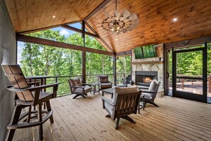 A wooden deck with outdoor seating, a fireplace, and large windows offering a view of trees. - Soaring Views - Mountain Cabin w/ all Kings, Hot Tub, Game Room, Fire Pit, Wi-Fi (Morganton)