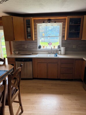 A kitchen with wooden cabinets, a stainless steel dishwasher, a window with a view of the outdoors, and a wooden dining table with chairs.