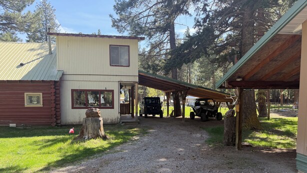 Soraa päällystetty ajotie johtaa rakennukseen, jossa on katettu kuisti. Rakennuksen vieressä on hirsihökkelö, ja kuistin alla on pysäköitynä ajoneuvo.