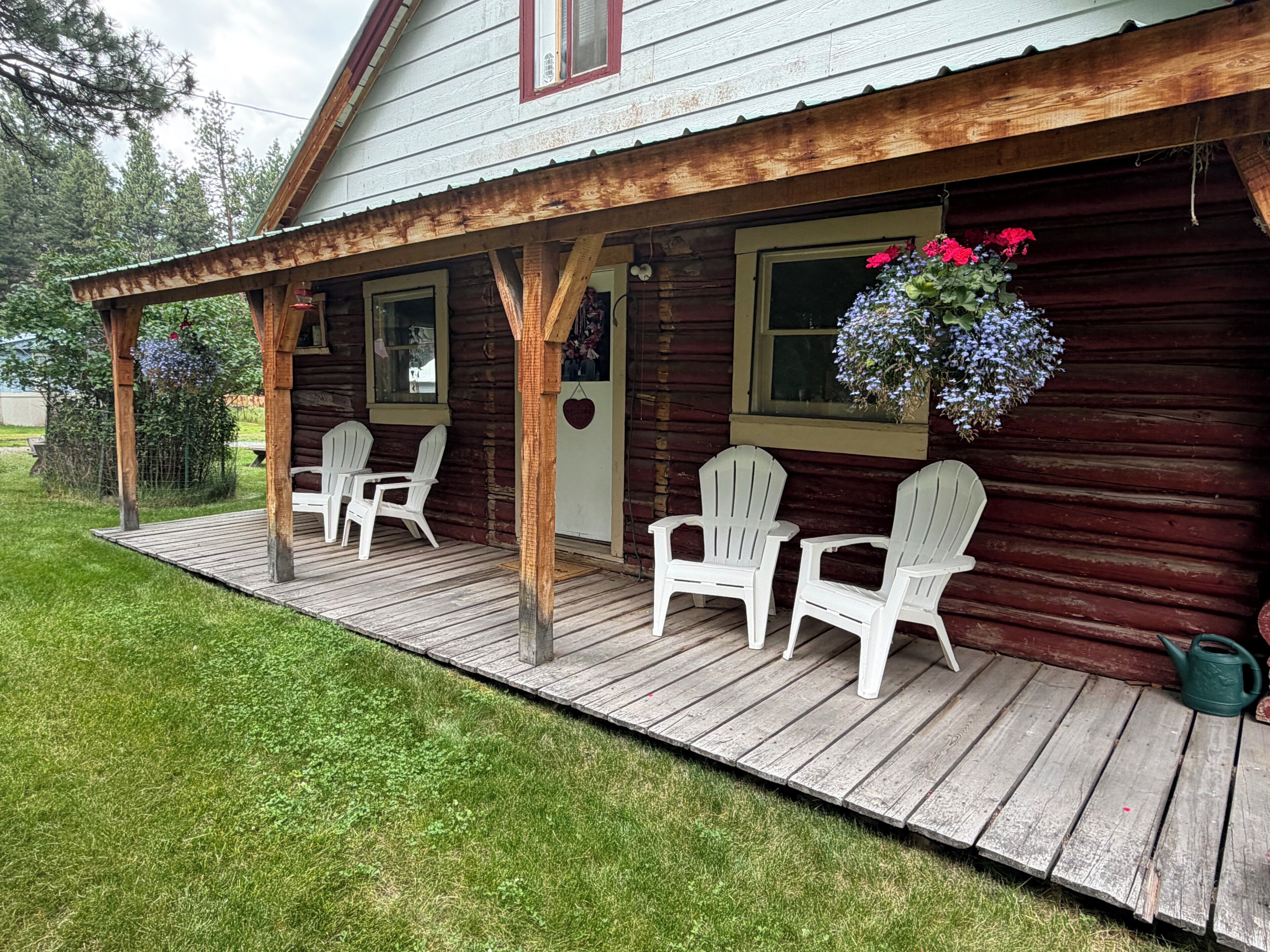 A wooden cabin with a porch, white Adirondack chairs, and a hanging flower basket.