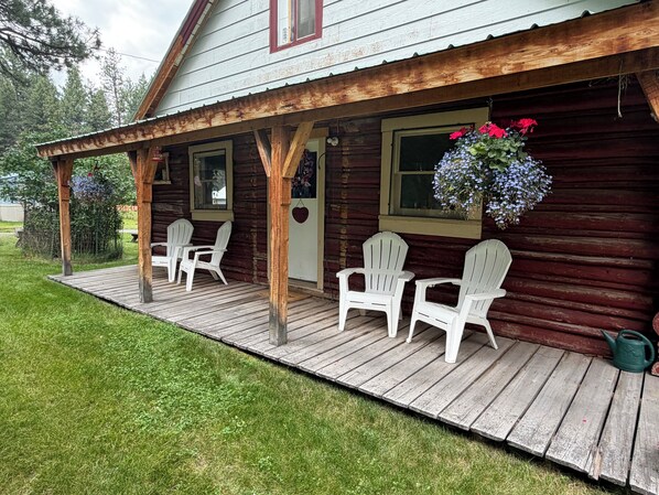 A wooden cabin with a porch, white Adirondack chairs, and a hanging flower basket.