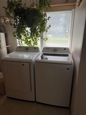 A laundry room with two white Samsung washing machines, a hanging plant, and a window with blinds. - Spacious 4-bedroom cabin in Lincoln perfect for your short or long term getaway! (Lincoln)