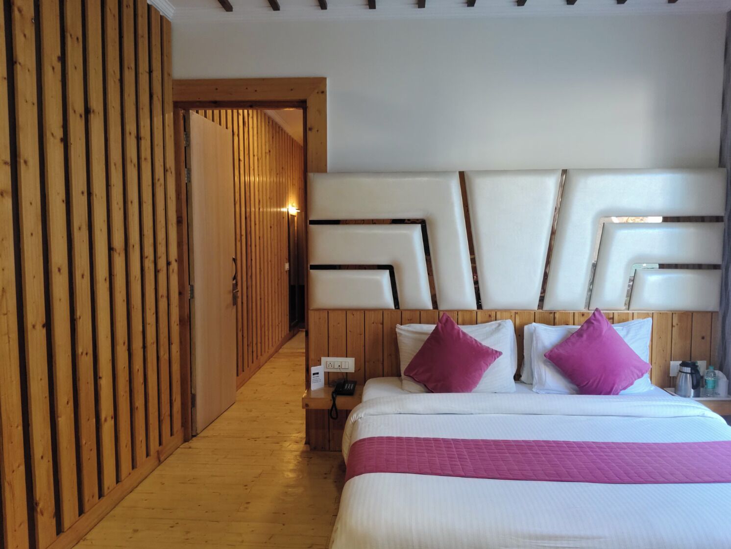 Een hotelkamer met een houten vloer, een bed met witte beddengoed en roze kussens, een houten hoofdbord en een houten scheidingswand.