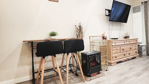 Un salon comprenant une table à manger en bois, deux chaises noires, un téléviseur fixé au mur, une commode en bois et un radiateur noir posé sur le sol.