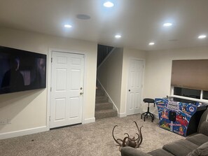 Ein Wohnzimmer mit an der Wand montiertem Fernseher, einer Treppe, einer Tür und einer Spielkonsole.