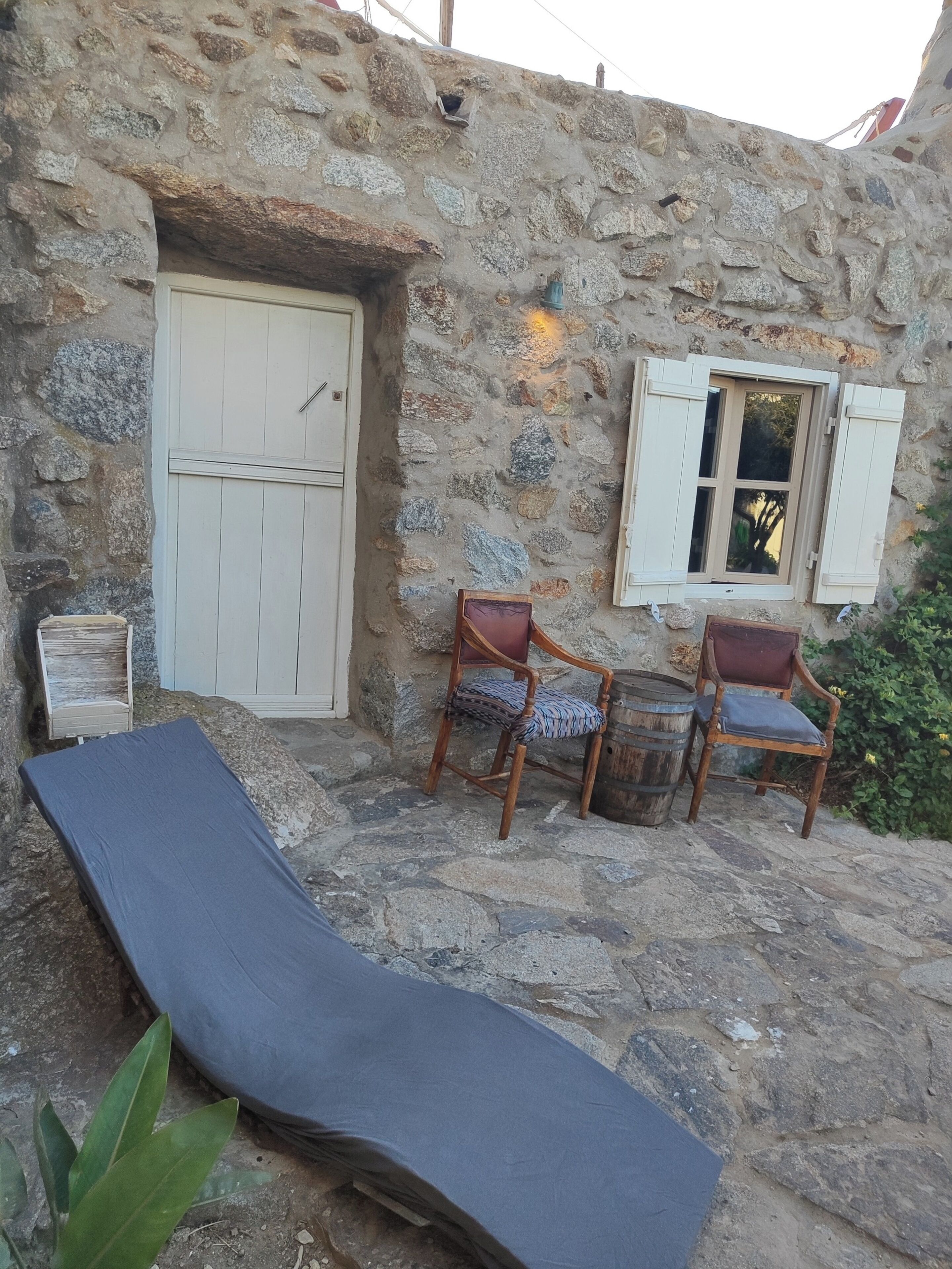 Un edificio de piedra con una puerta blanca, dos sillas de madera con cojines, una mesa pequeña y un tapete azul.