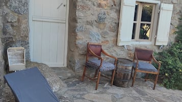 Un edificio de piedra con una puerta blanca, dos sillas de madera con cojines, una mesa pequeña y un tapete azul.