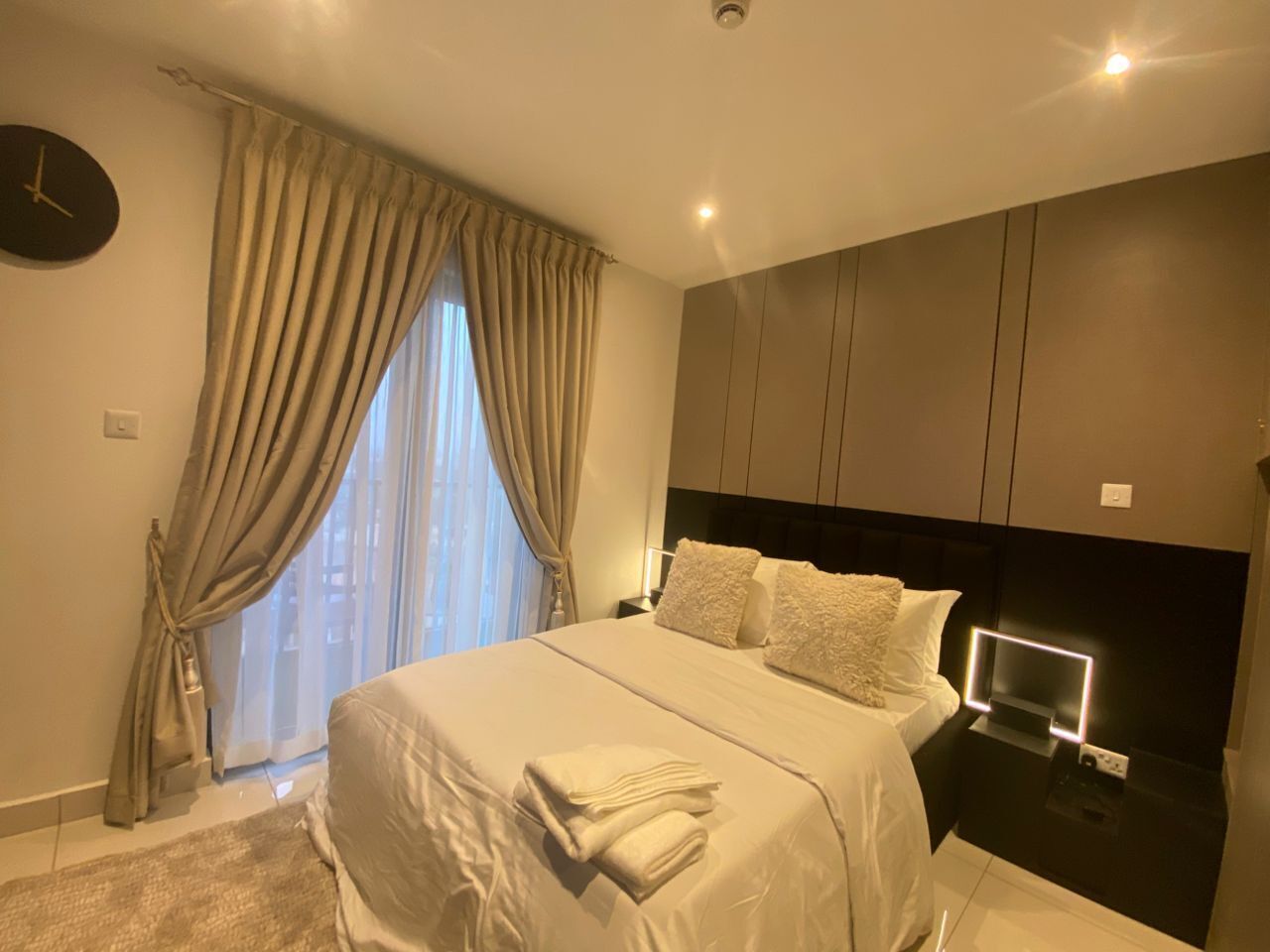 Un dormitorio moderno con cama, cortinas, un reloj y una mesita de noche con lámpara.