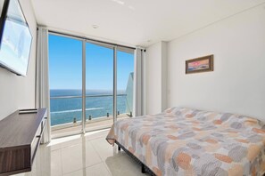 Ein Schlafzimmer mit Bett, Schreibtisch, Fernseher und einem großen Fenster mit Meerblick.
