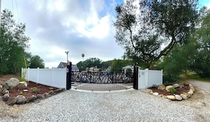 A gated entrance with a decorative metal gate, a white fence, and a gravel driveway. - Entire cozy two floor guest house located in rainbow near Temecula (Fallbrook)