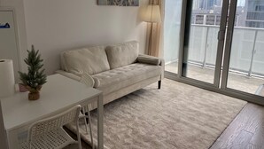 Ett modernt vardagsrum med en soffa, ett litet bord, en stol, en lampa och utsikt över staden genom ett stort fönster.