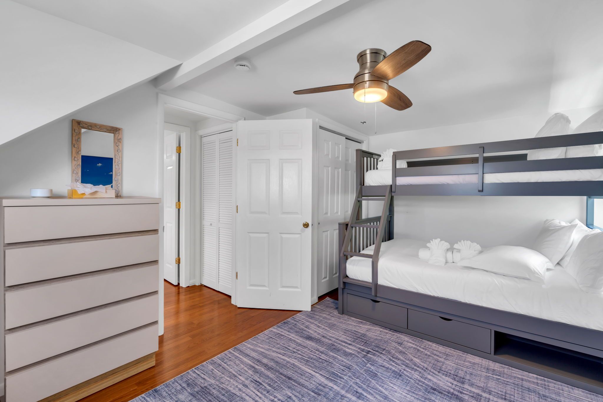 A bedroom with a bunk bed, a ceiling fan, a dresser, and a door leading to another room.