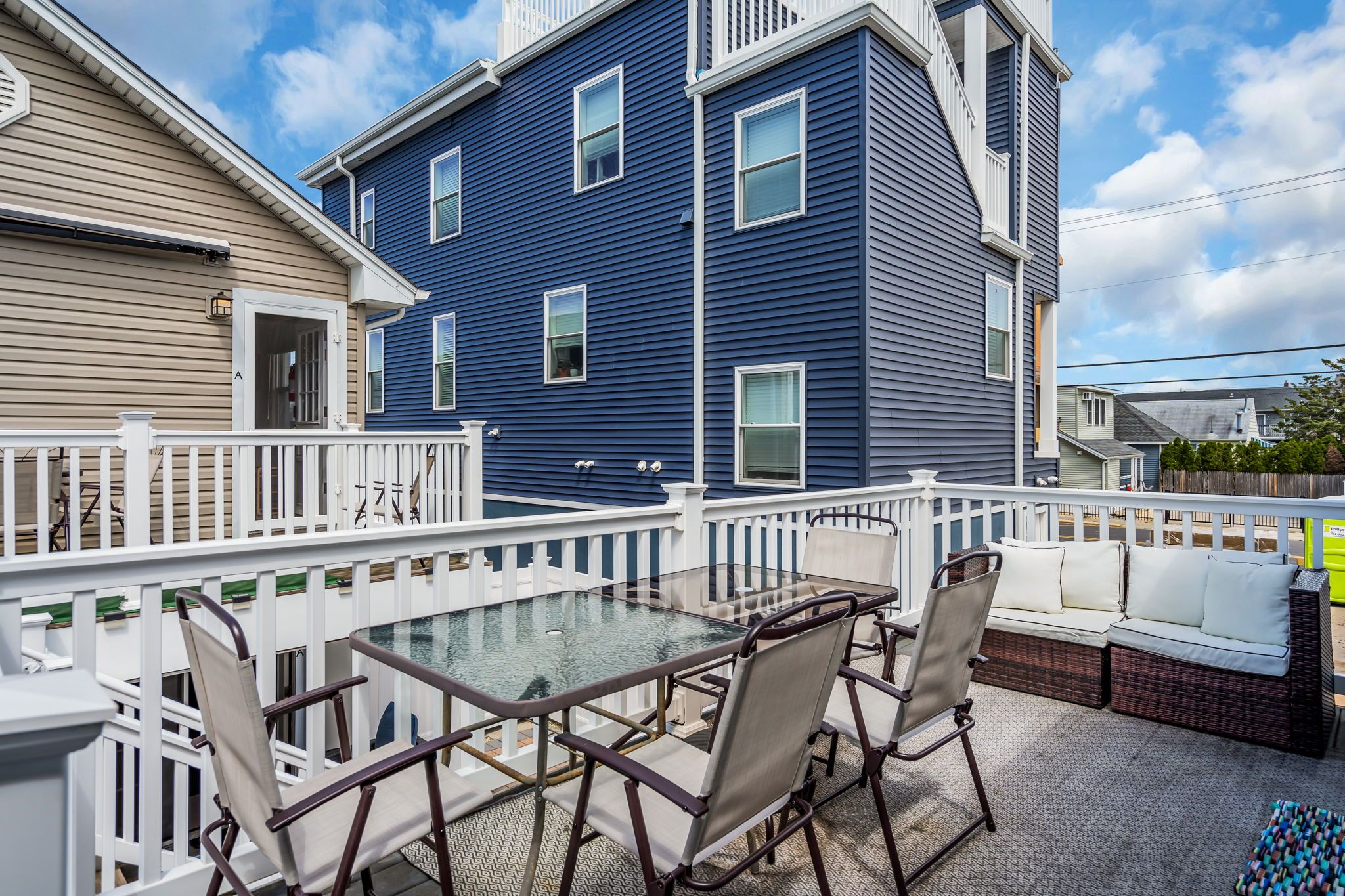 A balcony with a glass table and chairs, a white fence, and a blue building in the background.
