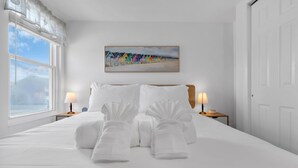 A hotel room with a bed, two bedside tables, a painting of beach huts, and a window with curtains. - Professionally Managed | 3 Blocks 2 Beach (Seaside Heights)