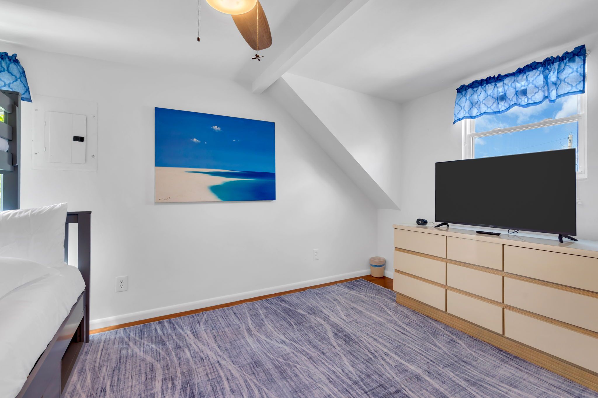 A bedroom with a bed, a TV on a stand, a painting of a beach, and a window with a view.