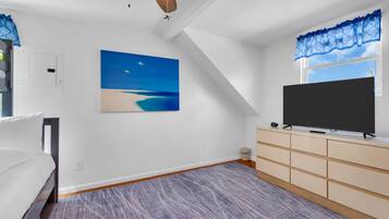A bedroom with a bed, a TV on a stand, a painting of a beach, and a window with a view.