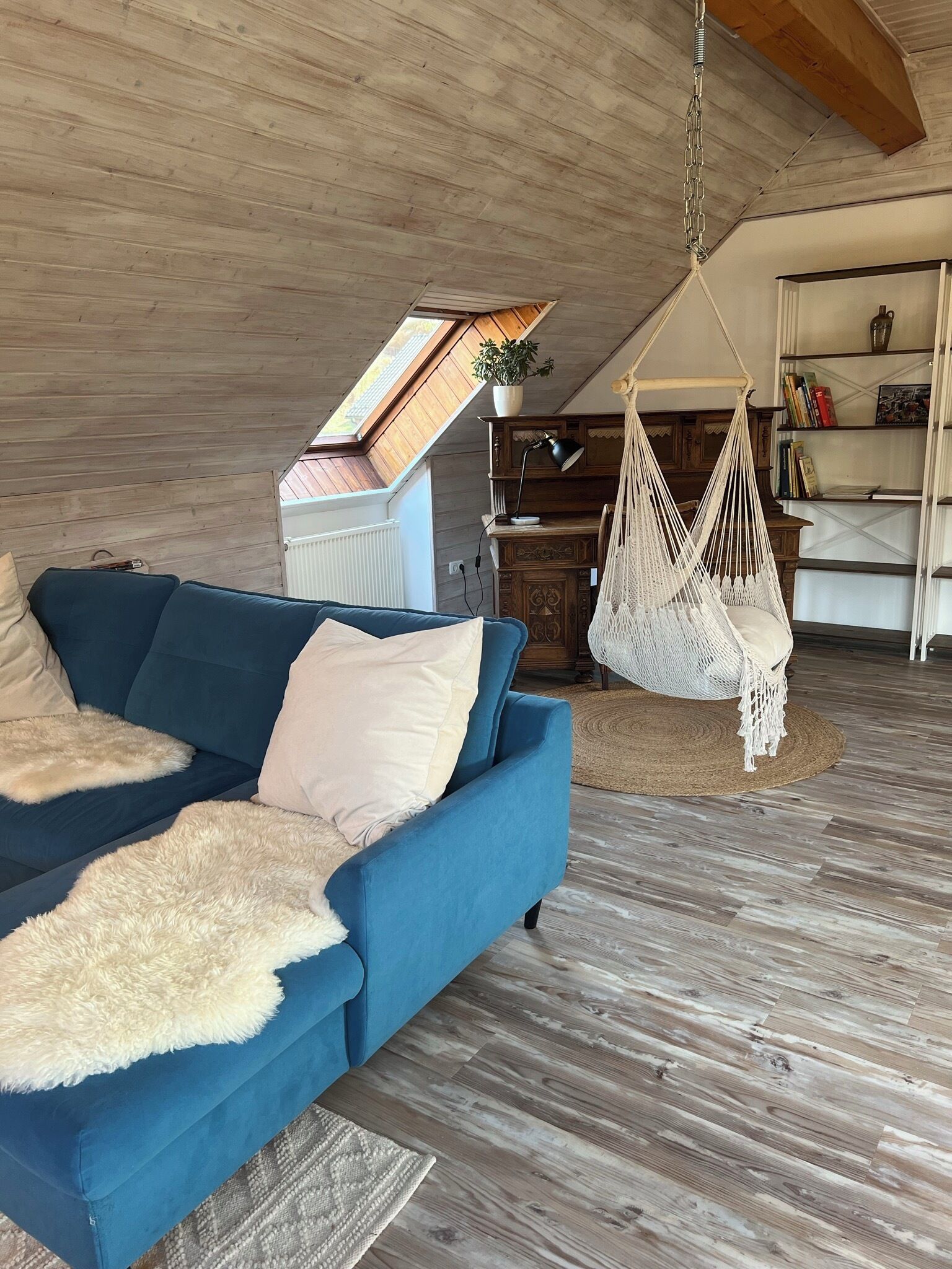 Ein gemütliches Wohnzimmer mit einem blauen Sofa, einem hängenden Makramee-Sessel, einem Holzregal und einem Dachfenster.