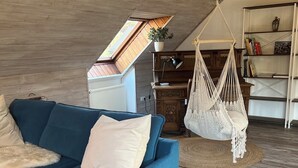 Un salon chaleureux avec un canapé bleu, un fauteuil suspendu en macramé, une bibliothèque en bois et une lucarne.