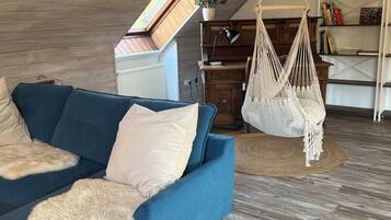 Ein gemütliches Wohnzimmer mit einem blauen Sofa, einem hängenden Makramee-Sessel, einem Holzregal und einem Dachfenster.