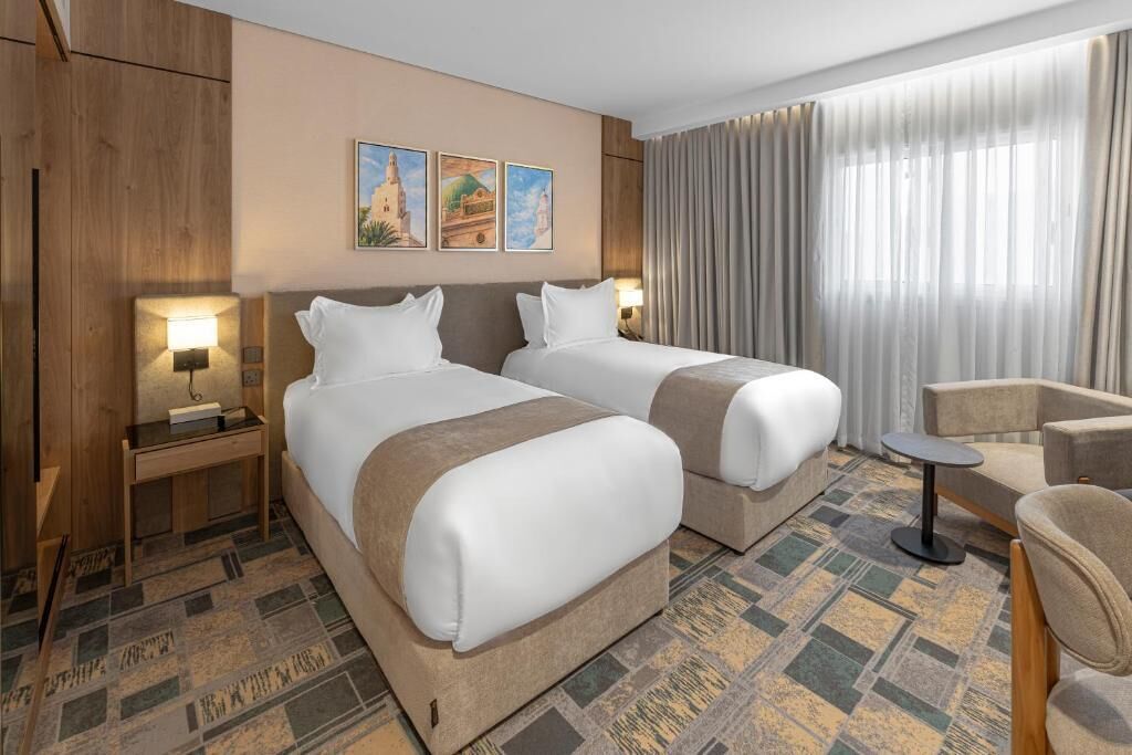 Habitación de hotel con dos camas, un escritorio pequeño, una silla y un ventanal con cortinas.