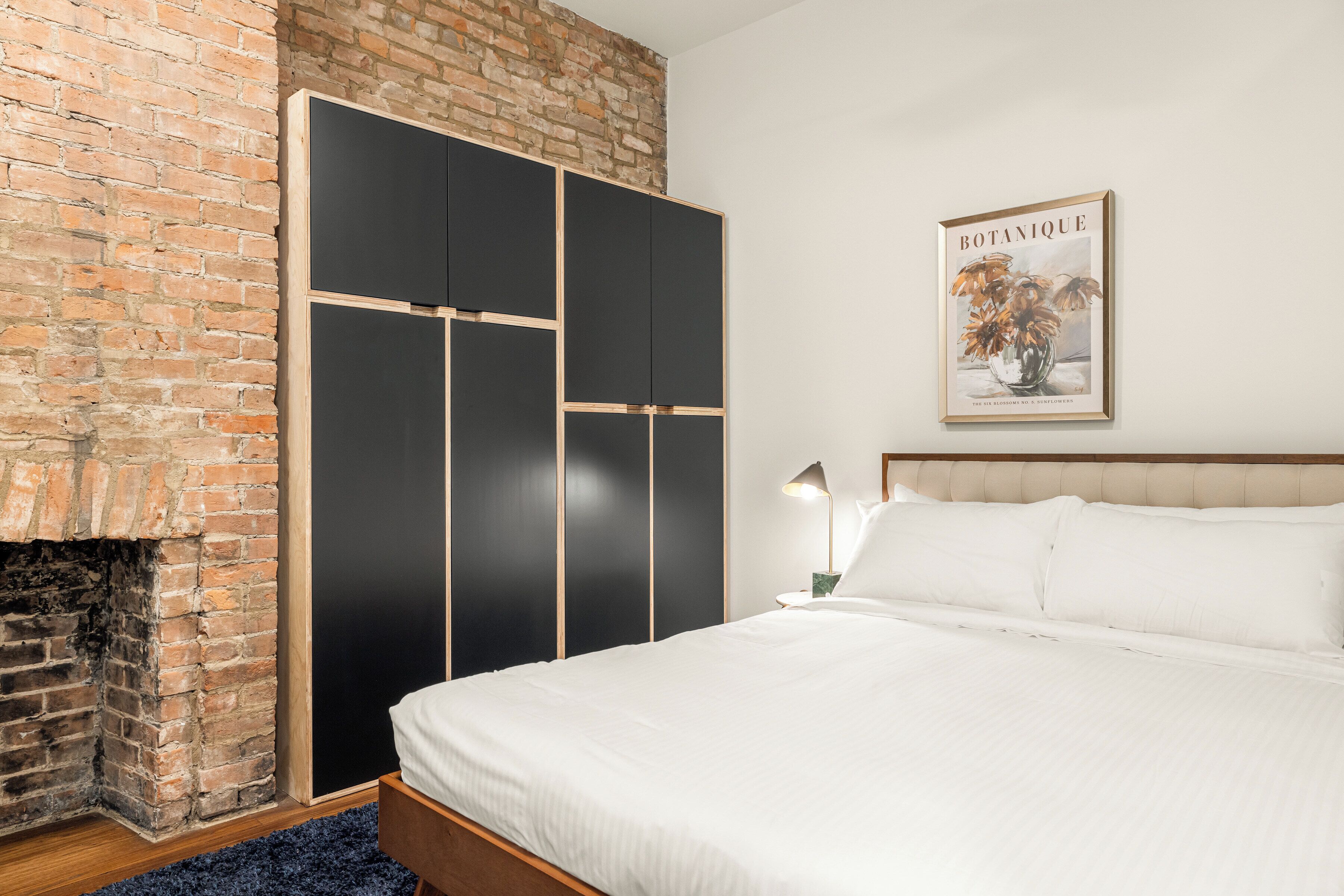 A bedroom with a brick fireplace, a black wardrobe, a bed with white bedding, and a framed botanical print on the wall.