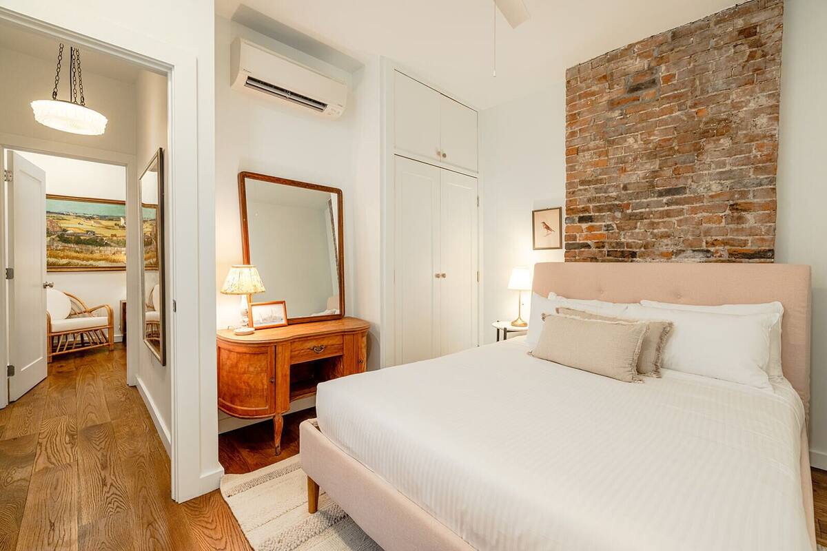 Un dormitorio con una cama grande, un aparador de madera, un espejo, una pared de ladrillo y una puerta que da a otra habitación.