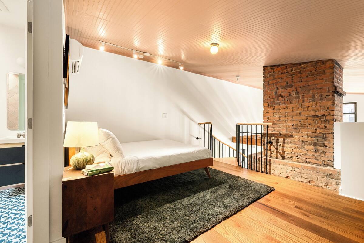 Una habitación con una cama, una mesita de noche de madera, una alfombra verde, una pared de ladrillo y una escalera.