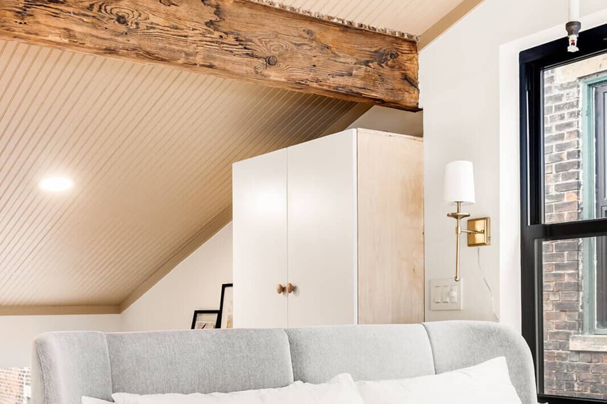 Un dormitorio con cama, techo de madera, ventana y una lámpara fijada a la pared.