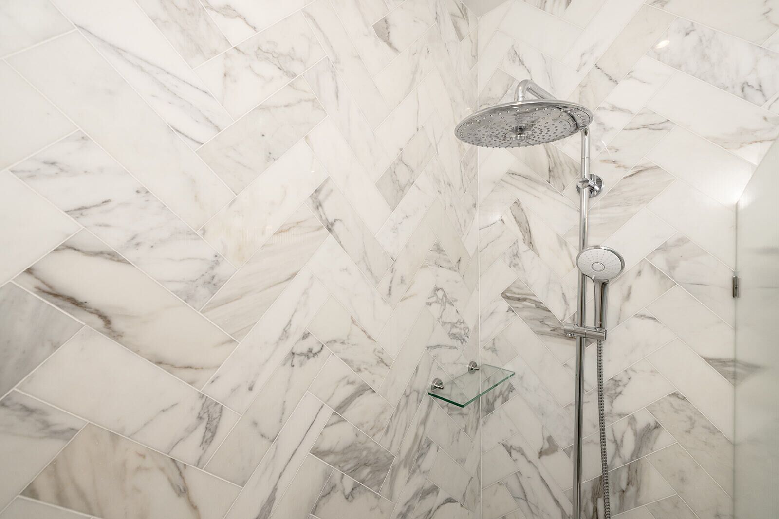 A shower with a rain showerhead and a handheld showerhead, set against a herringbone-patterned marble wall.
