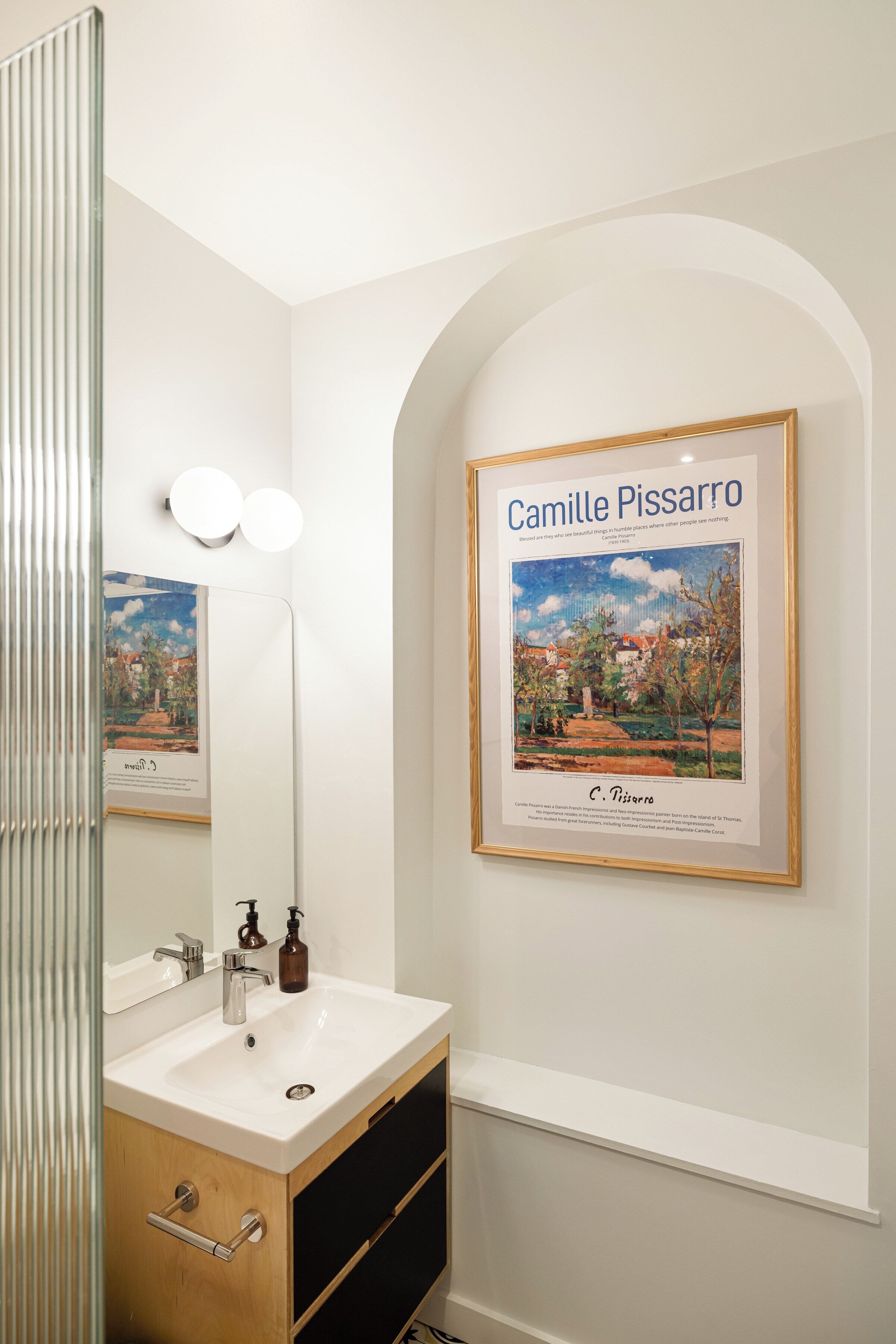 A bathroom with a white sink, mirror, and framed artwork of Camille Pissarro.