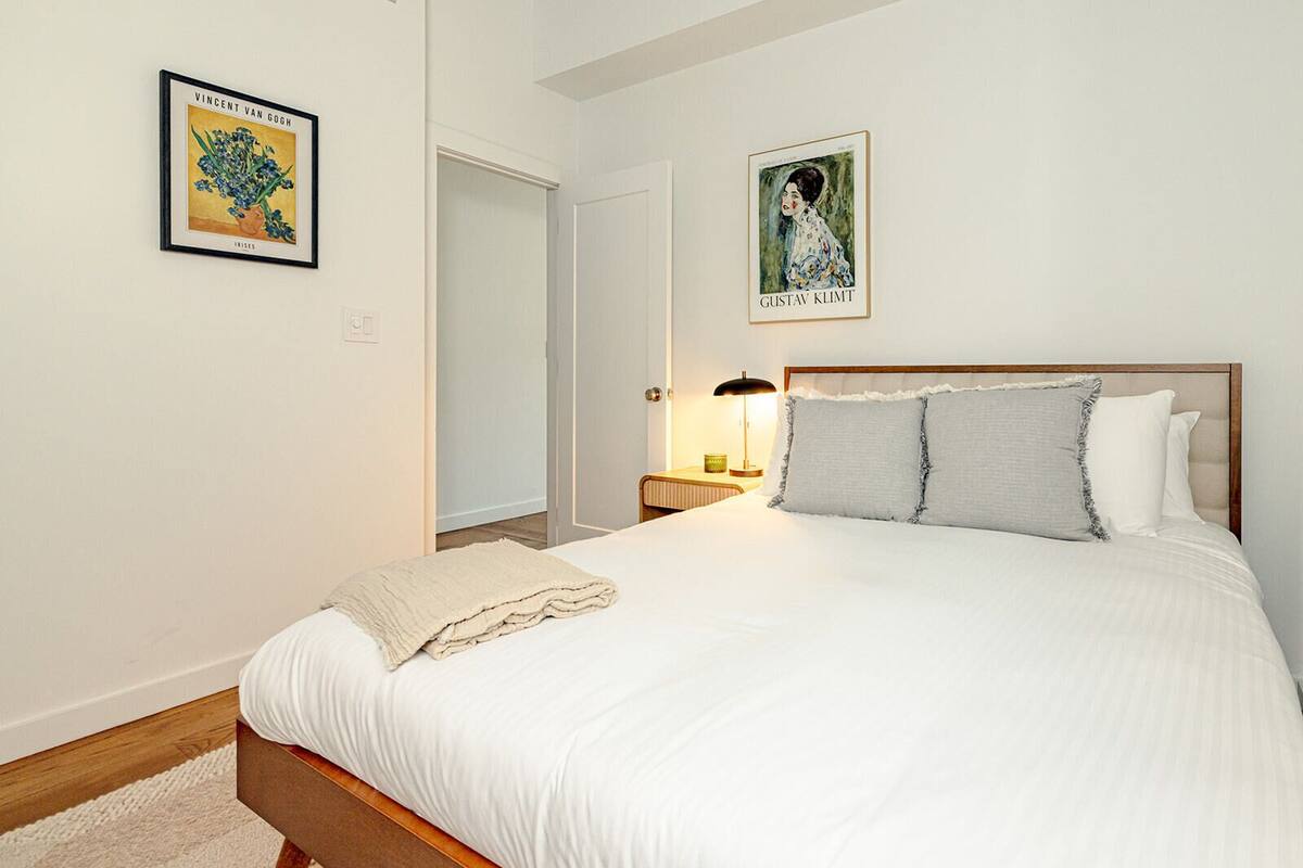 Un dormitorio con cama, mesita de noche, lámpara y dos cuadros en la pared.