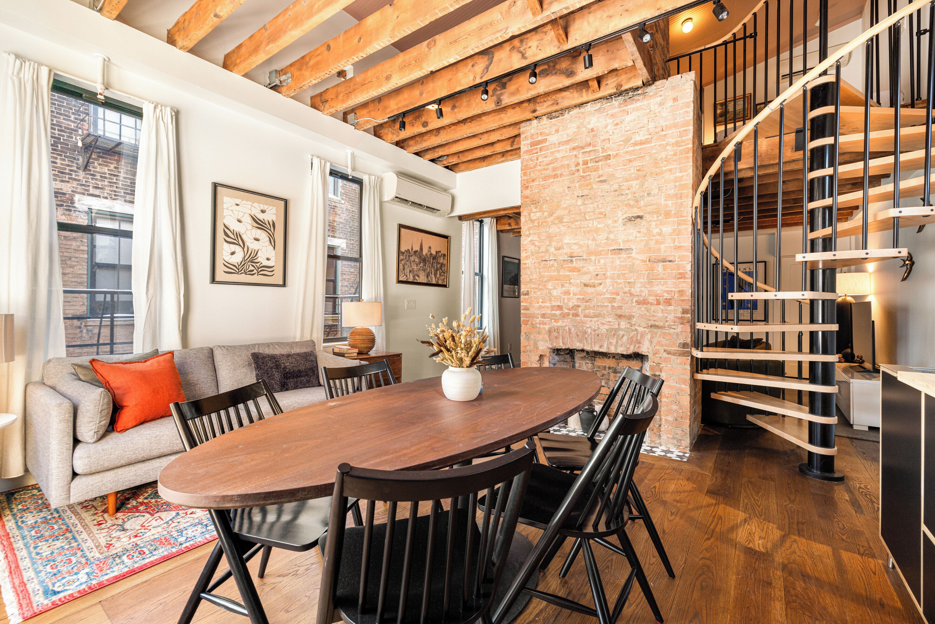 A spacious living area with a wooden dining table, comfortable seating, a brick wall, and a spiral staircase.