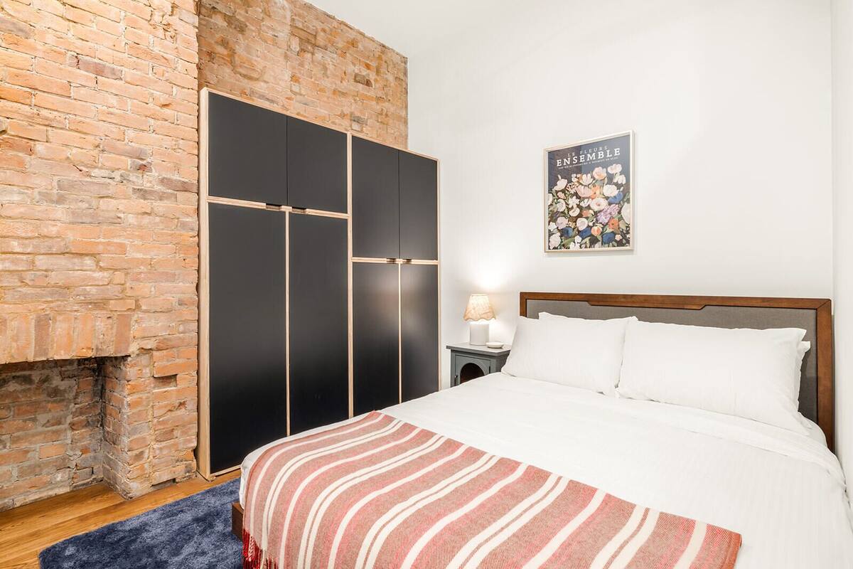Un dormitorio con pared de ladrillo, una cama con ropa blanca, una manta a rayas, una mesita de noche con una lámpara y un cartel en la pared.