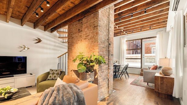 A modern living room with a brick wall, wooden beams, a flat-screen TV, a sofa, a coffee table, and a dining area with chairs.
