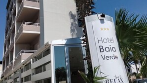 Monikerroksinen hotellirakennus, jossa on parvekkeita, kyltti, jossa lukee ”Hotel Baia Verde”, ja tunnuslause: ”Nyt on aika rentoutua”.