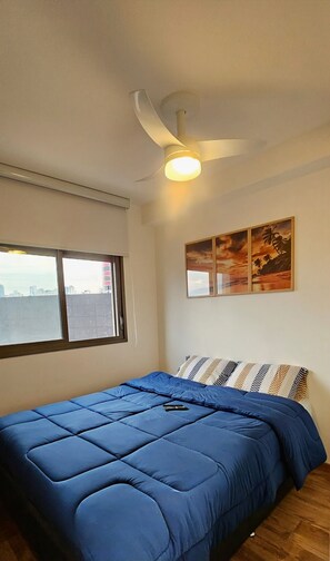 Une chambre à coucher avec un lit, un ventilateur de plafond, une fenêtre et un mur orné d’un tableau.